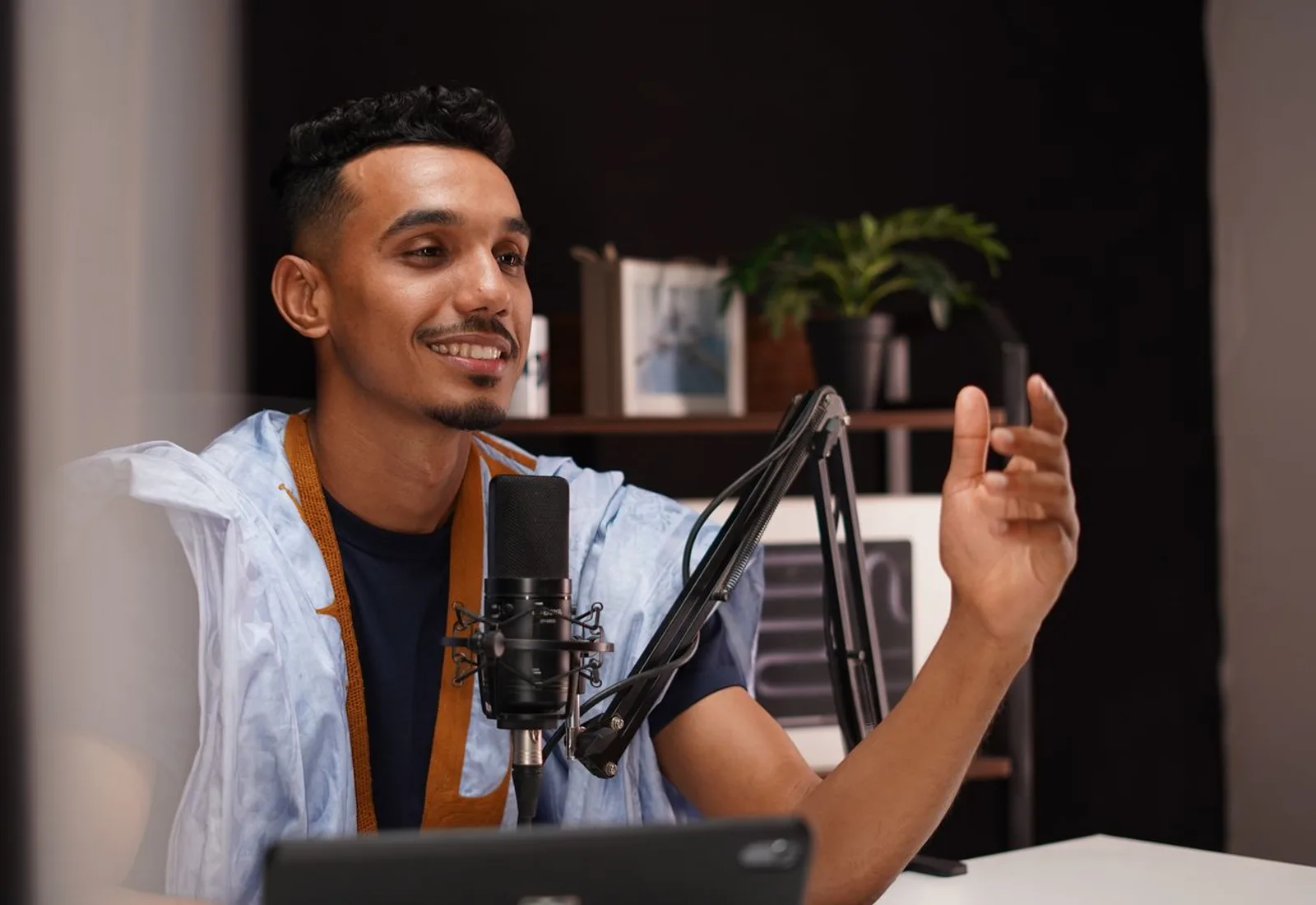 A creator mid-record at his podcast desk, smiling and gesturing into the mic